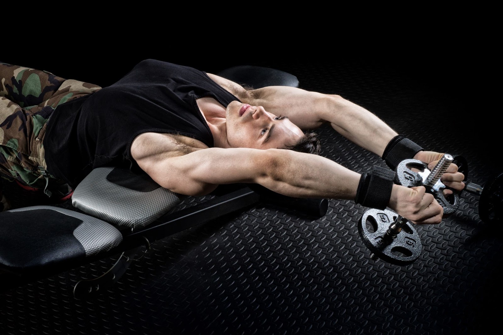 Young muscular man in a black tank top performing a dumbbell pullover pull workout.