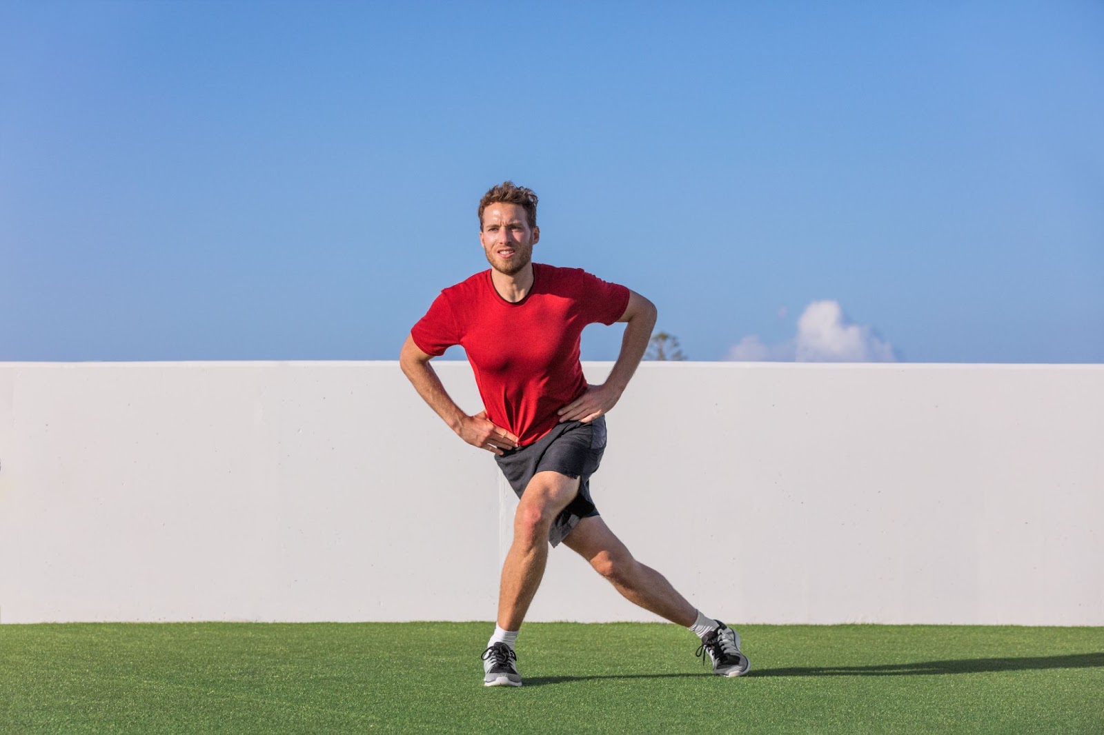 Fit man in red shirt, performing a curtsy lunge outside on astro turf