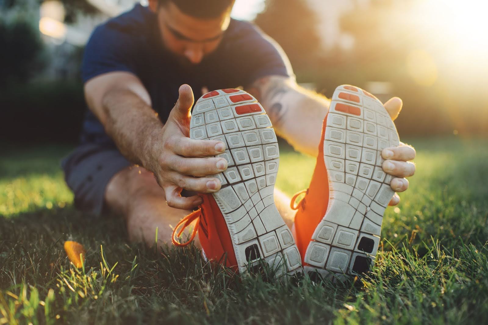 Flexible man extending his back and holding his feet, while sitting on grass